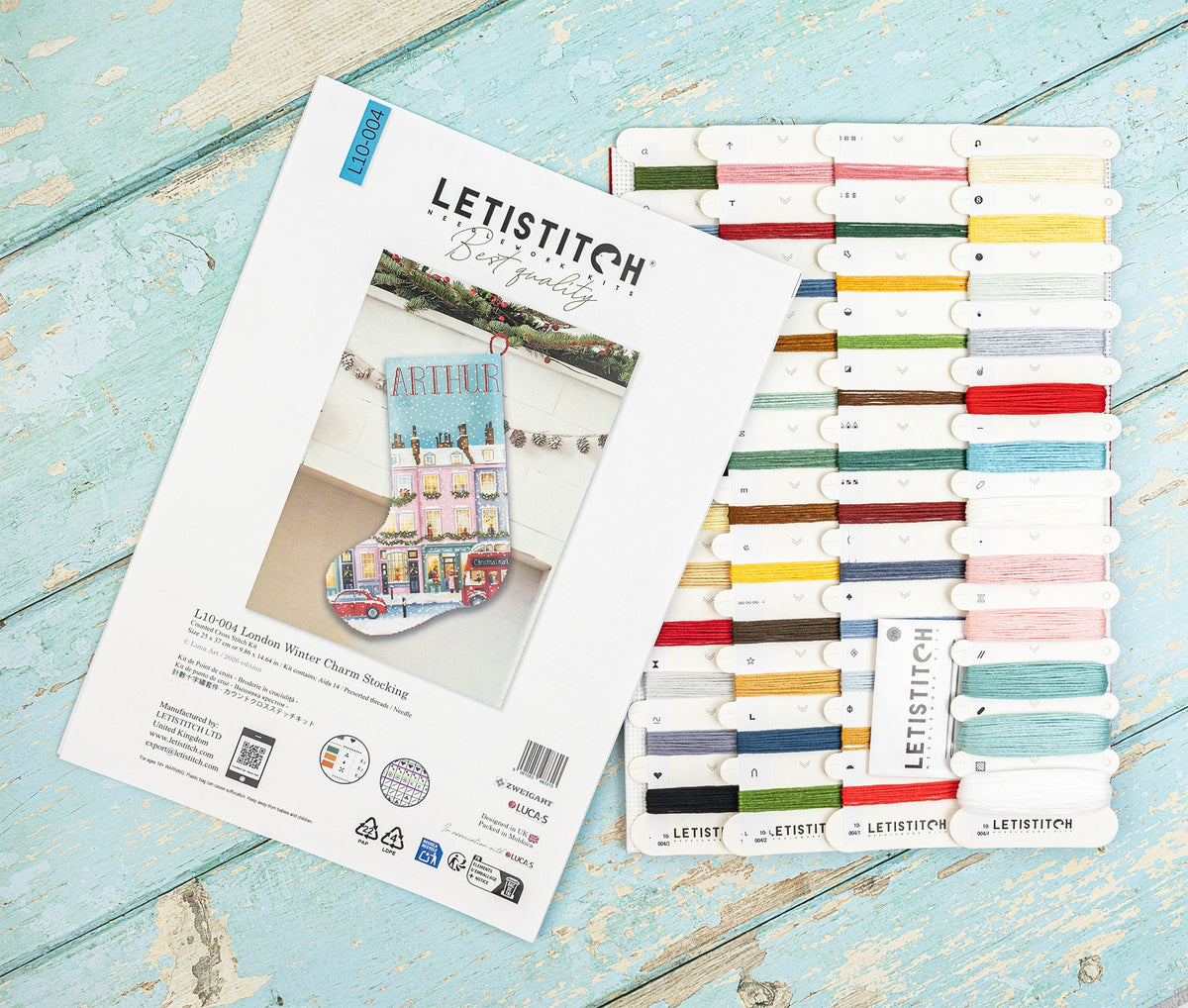 A flat-lay of the Letistitch London Winter Stocking kit, showing the printed instruction booklet next to a rack of colorful embroidery floss threads on a distressed light blue wooden background.