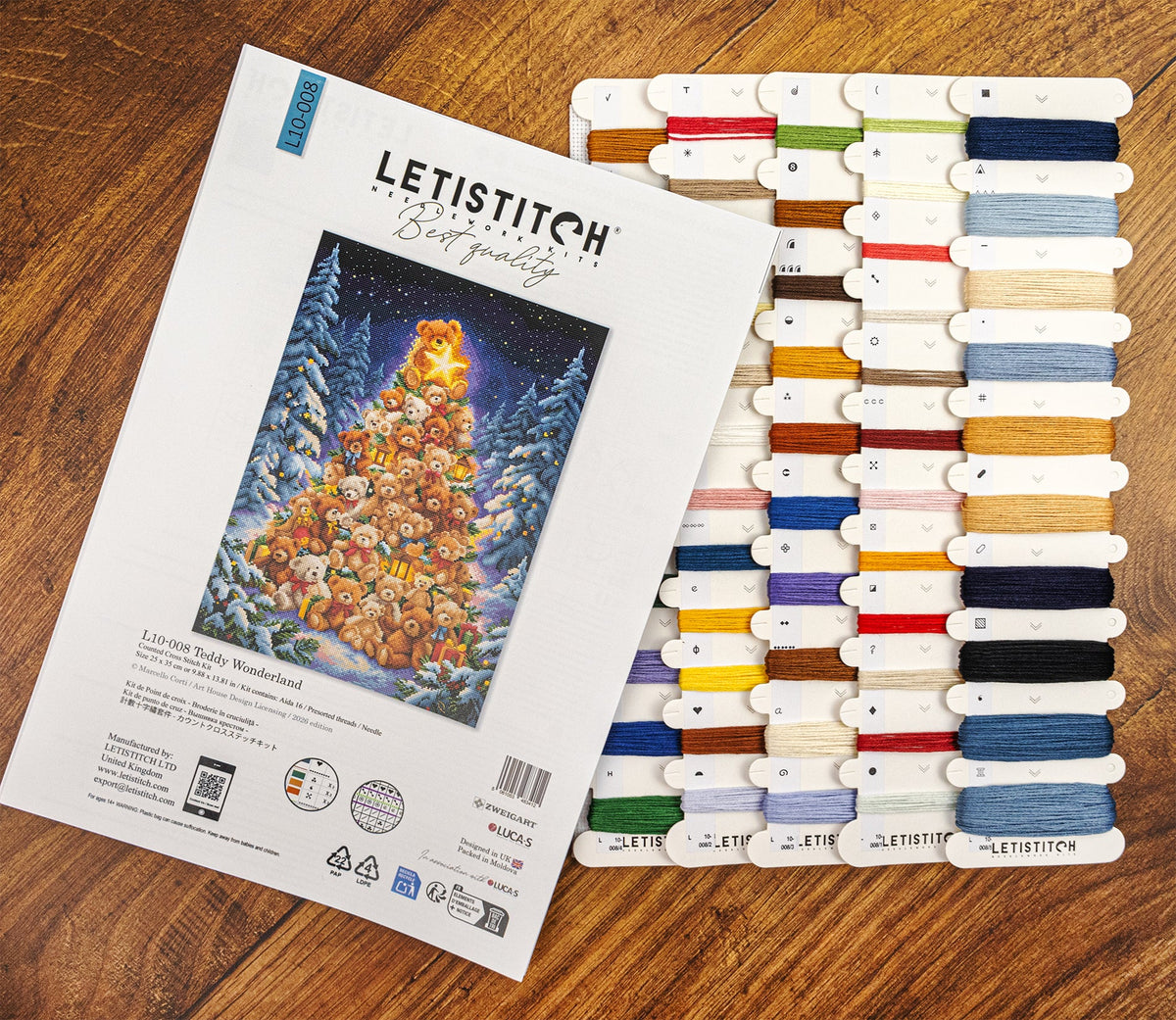 A Letistitch Christmas Tree cross-stitch kit shown with its printed cover and a large array of colorful thread spools arranged on a wooden surface.
