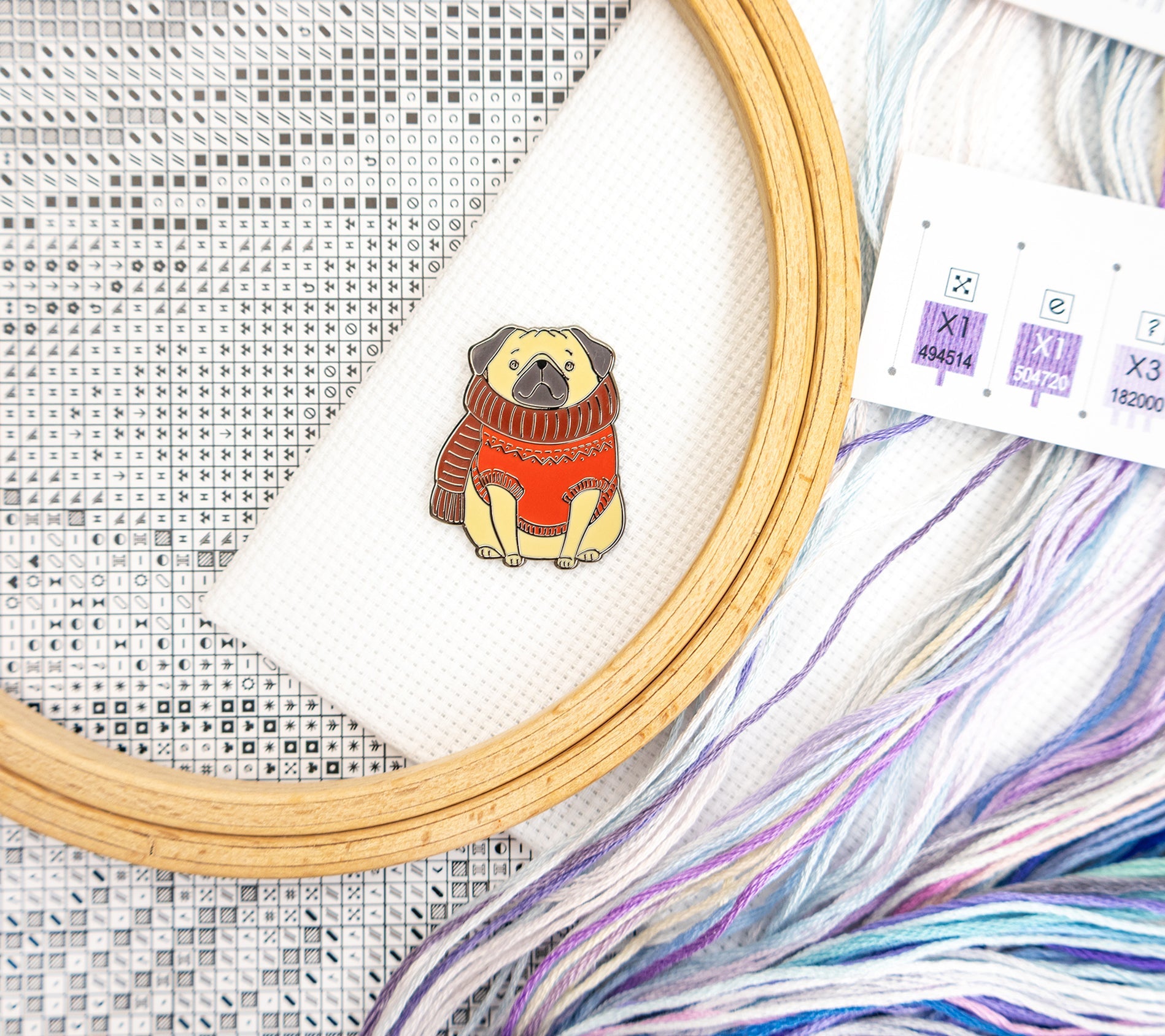 Embroidery hoop with a pug wearing a sweater design, surrounded by embroidery threads and fabric.
