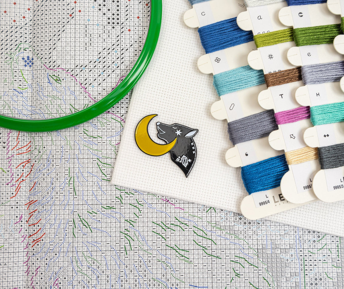 A close-up flat lay of a cross-stitch kit or craft setup on white canvas, including a green hoop, a small wolf-and-moon enamel pin, thread spools, and a counted fabric chart.