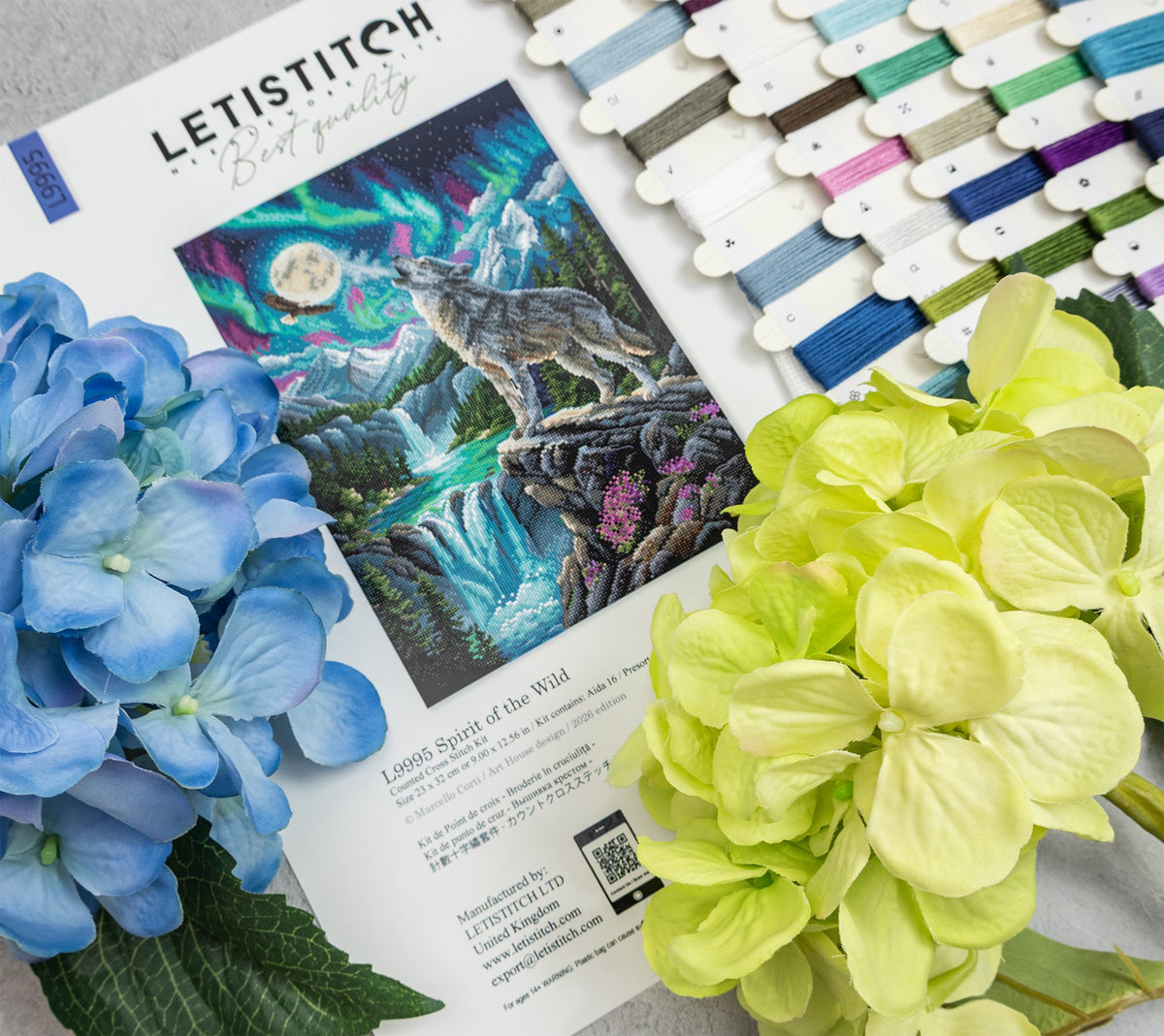 A flat lay of a Letistitch embroidery kit showing the pattern booklet, a row of colorful thread spools, and decorative hydrangea flowers on a light gray surface.