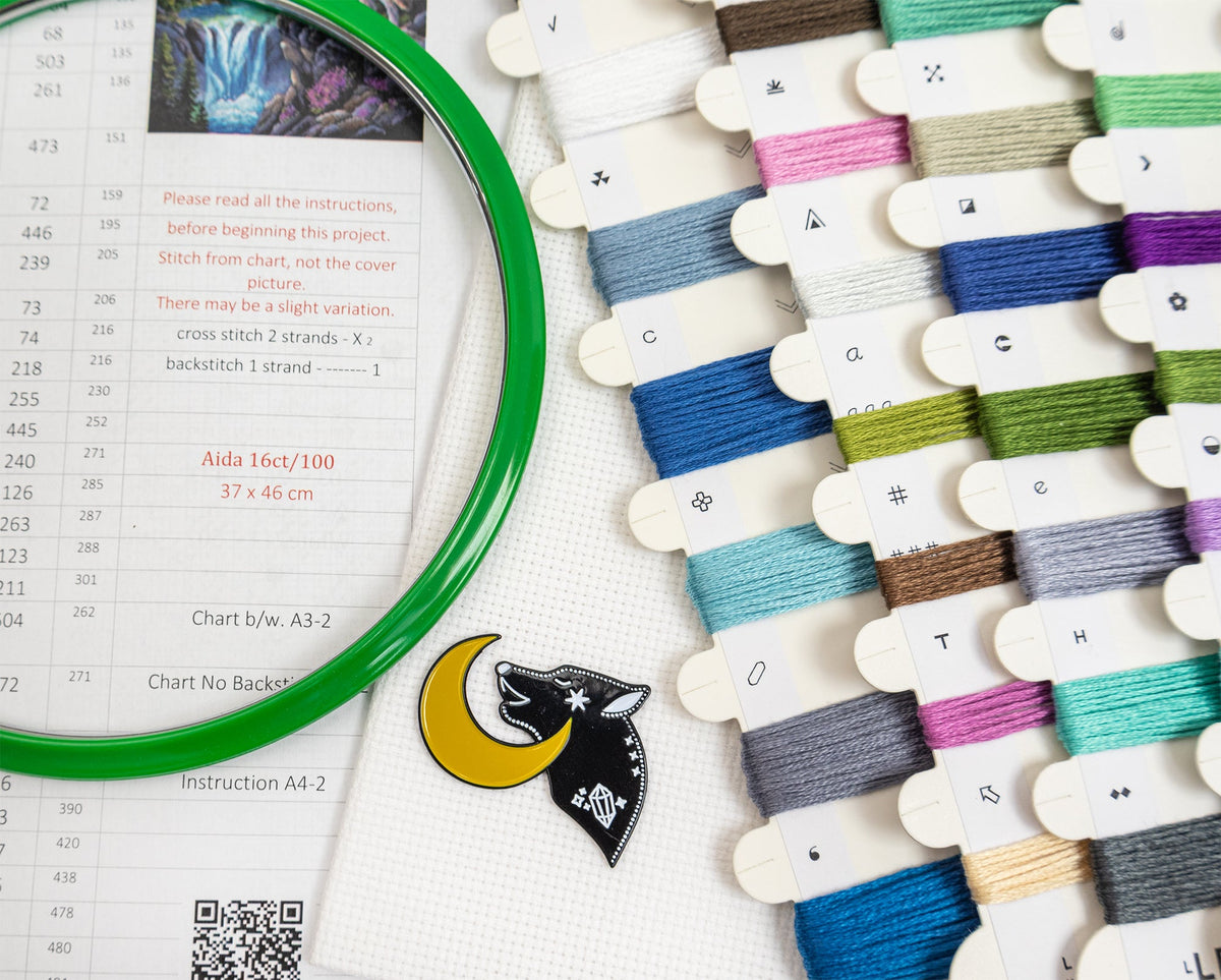 A close-up of a sewing or embroidery instruction sheet with a green circular magnifier, a small wolf emblem, and multiple thread spools in assorted colors arranged on a white surface.