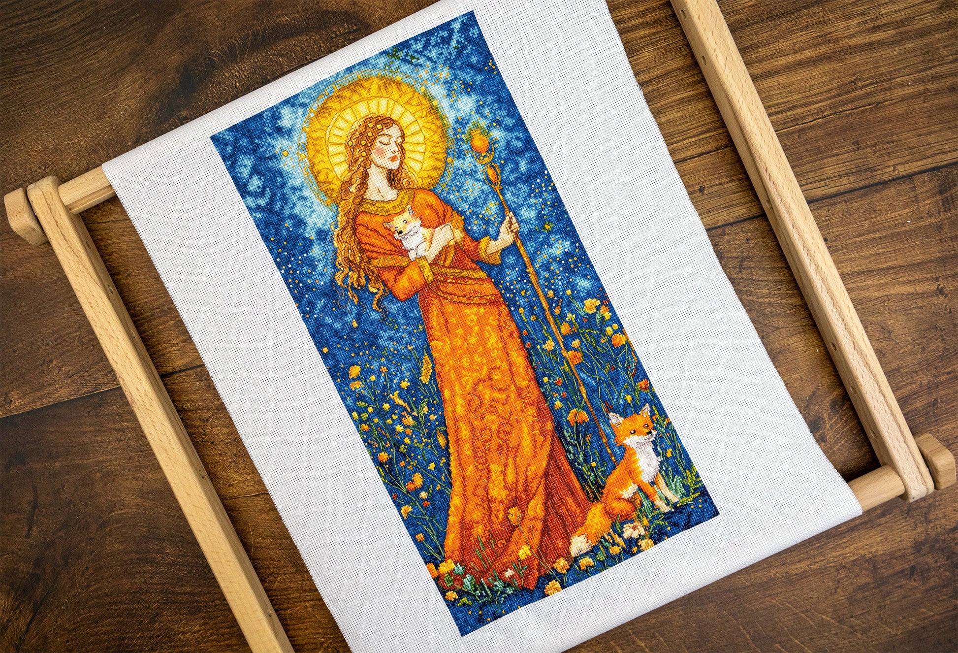 Cross-stitched piece of art on fabric with a woman holding a staff and a dog, framed in wooden hoop on a wooden surface.